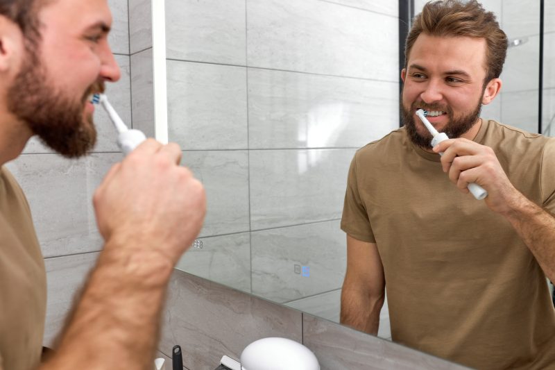 man looking in mirror while brushing teeth with electric toothbrush